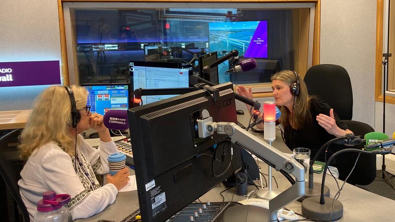 Two women sitting opposite one another in a radio studio talking into microphones with computer monitors around them 