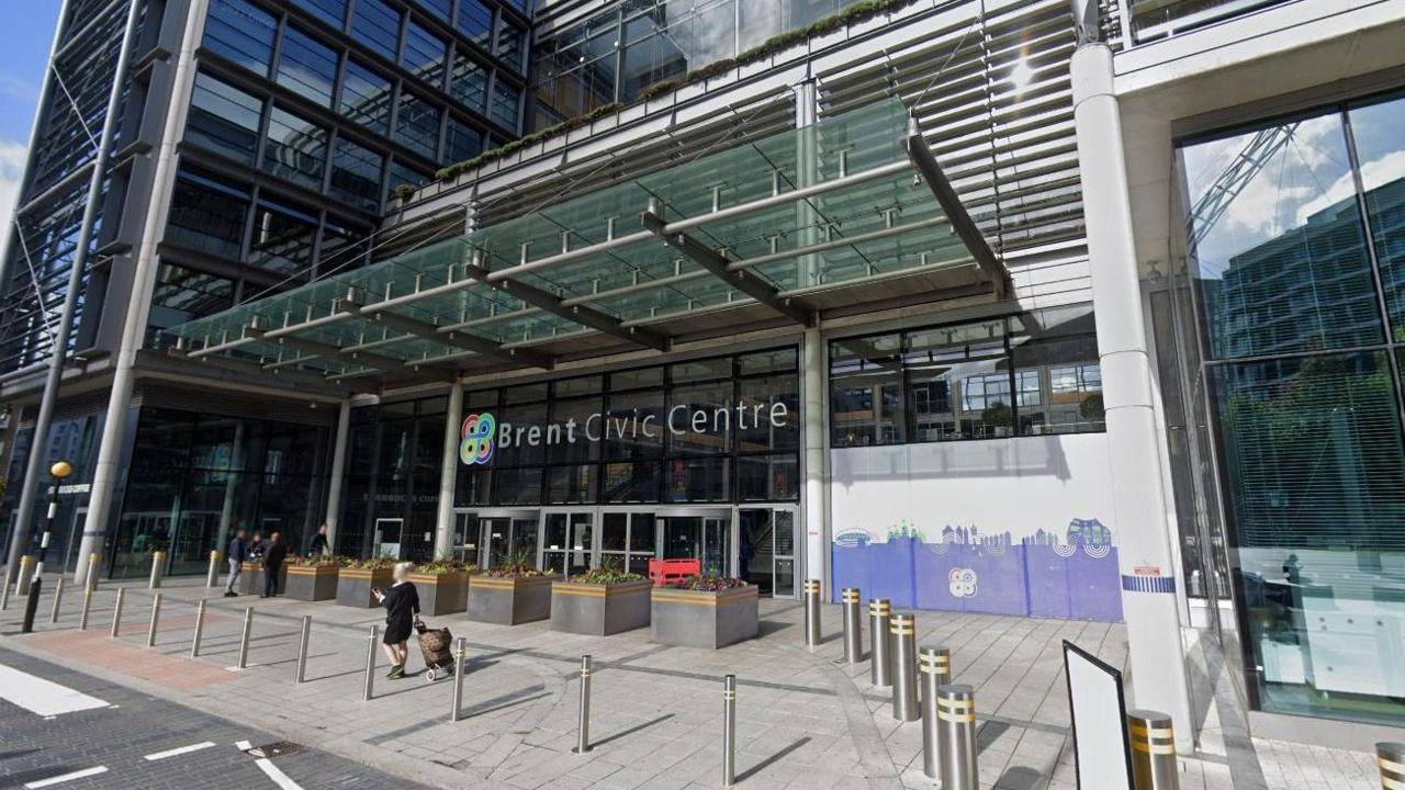 A general view of the outside of Brent Civic Centre. The Brent Council logo is in view alongside the writing of the name of the centre. There are people that can be seen in the image also.