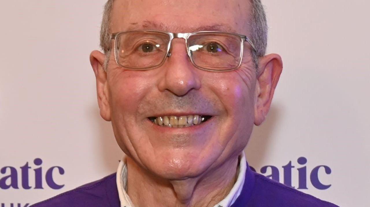 A man in a purple jumper wearing rectangular glasses smiles at the camera