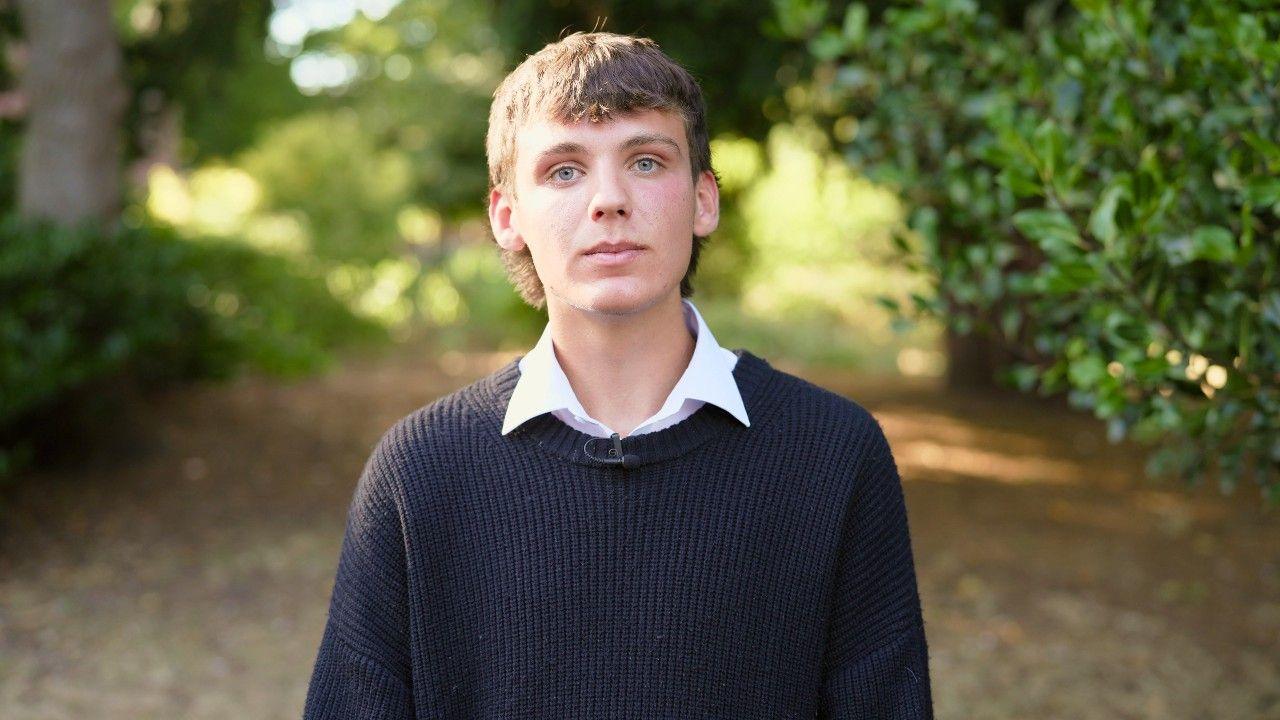 A young man with blond hair and wearing a dark jumper stands on a path in a forest