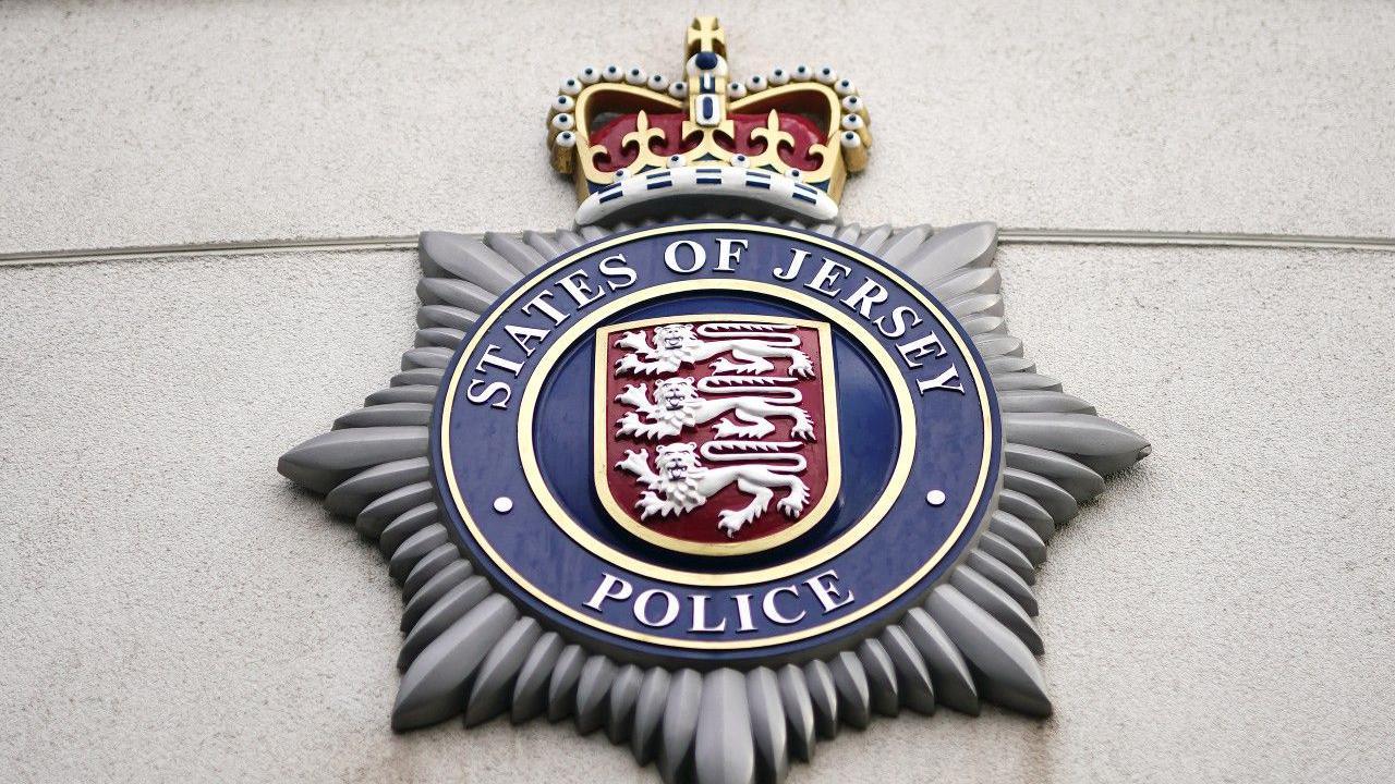The States of Jersey Police crest mounted on a wall. It is shaped like a septagram within which is a circle holding the Jersey shield featuring three lions and the words STATES OF JERSEY POLICE around the outside. There is a crown at the top of the crest.