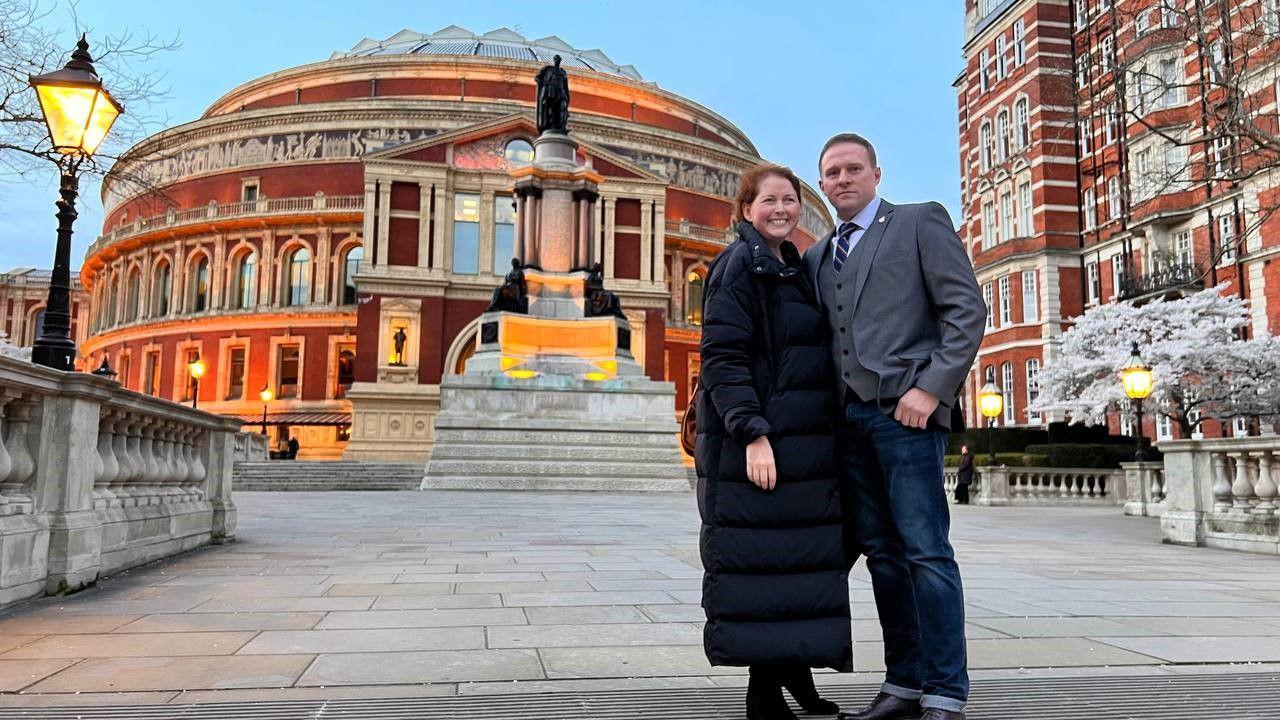 A man wearing a suit stands next to a woman in a full body coat. A street lamp is lit to their left. Flagstones lead to a black statue behind them and a domed brick building.