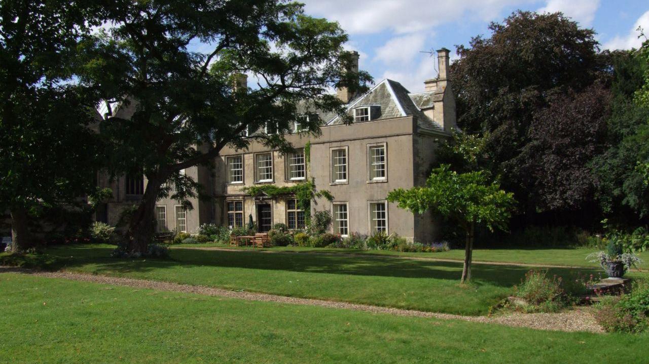 The two storey house has large white windows and a third tier in the roof.  The second photo is taken from a slightly different angle and shows the trees in the gardens.