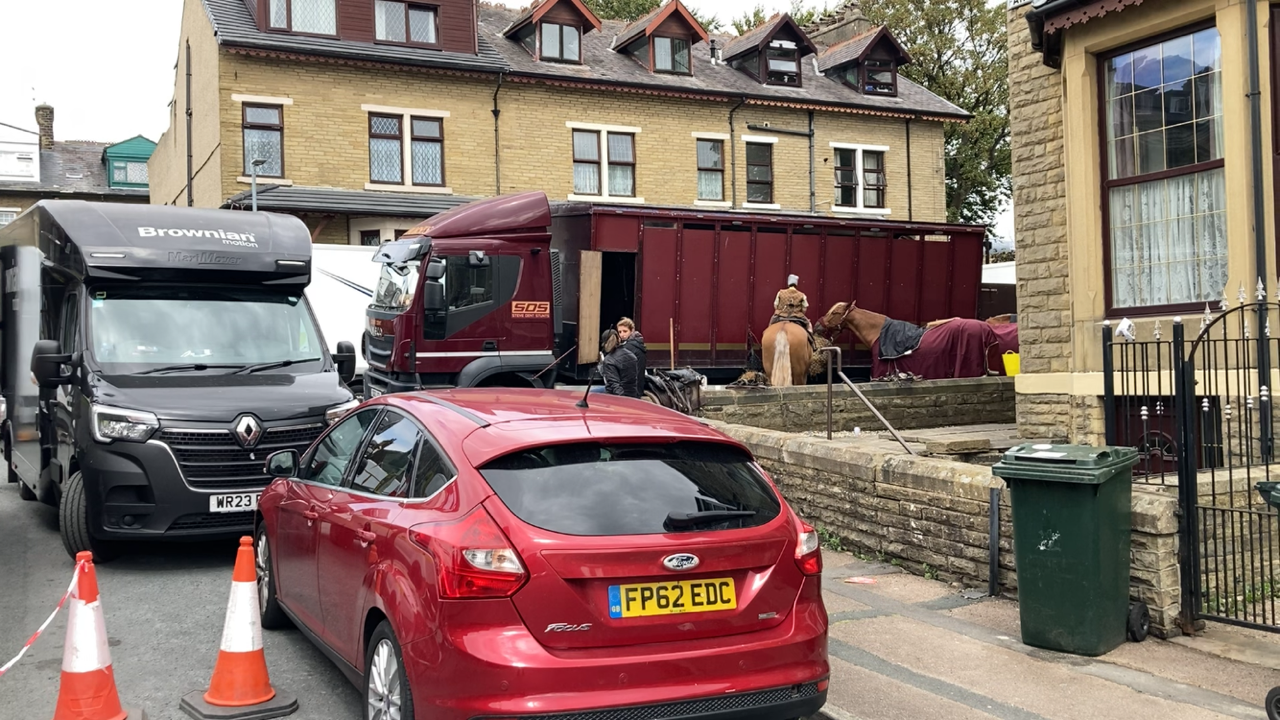 A car parked on a street with horseboxes in the background with a horse tethered to its side.