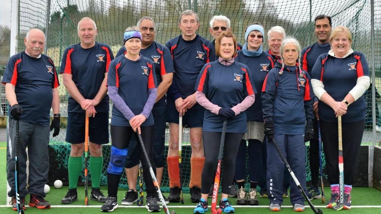 A group of 12 members of the hockey team, all wearing their uniforms and holding hockey sticks, they are standing in front of a goal