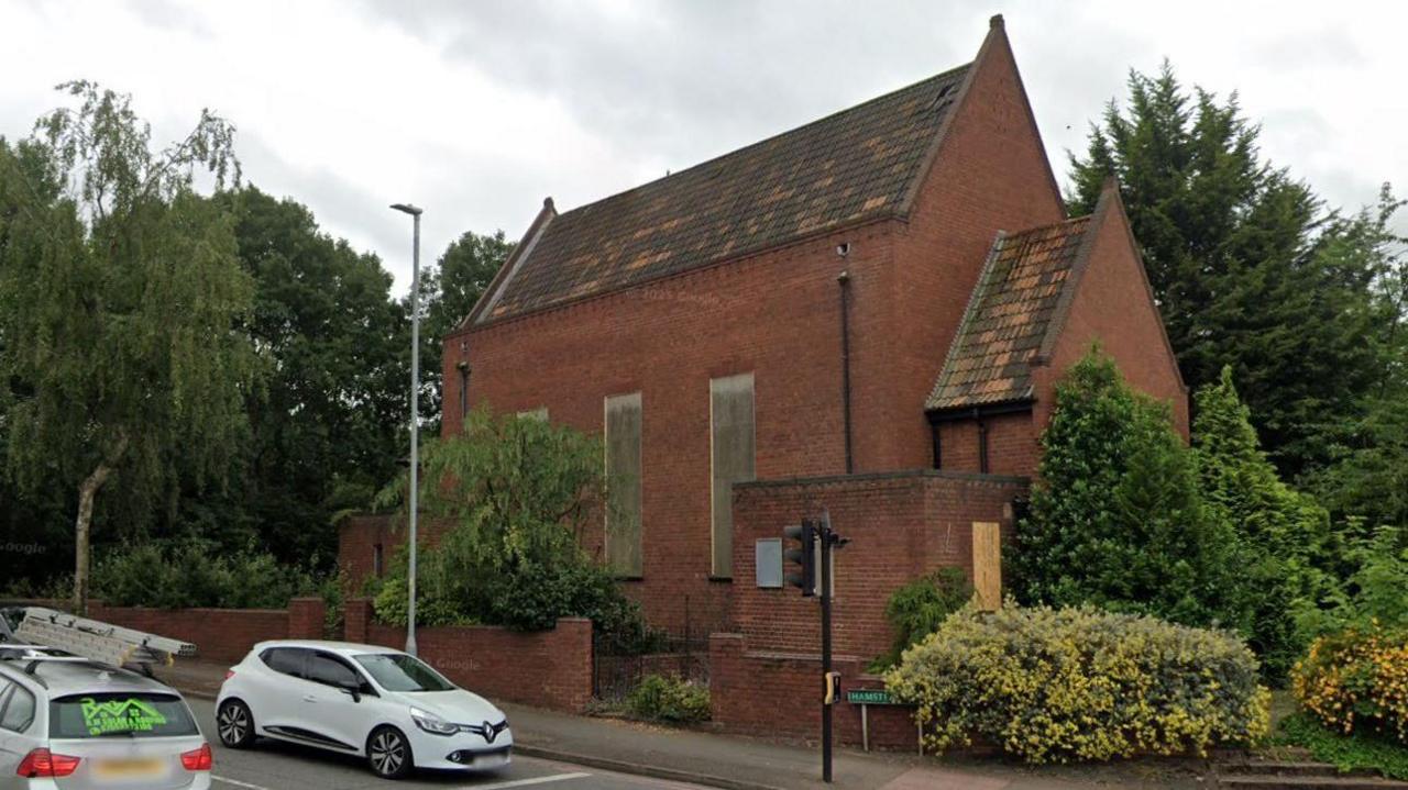 A boarded up church building stands on the corner of Hamstead Road. There are cars on the road, and there are shrubs and trees planted around the church. The building has a steep pointed roof and drainpipes on the sides.
