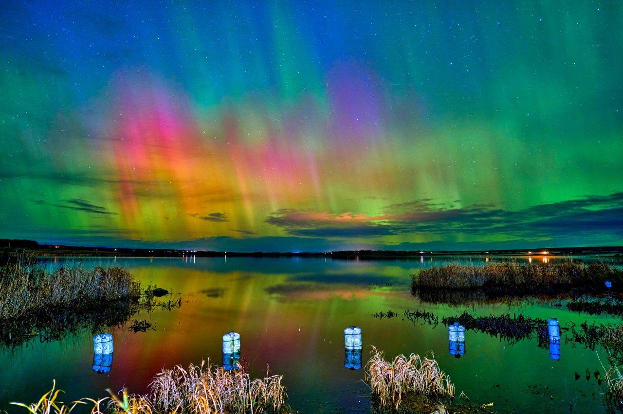 The aurora, which has green, pink, yellow and blue colours, shining over a loch with lanterns in the foreground.