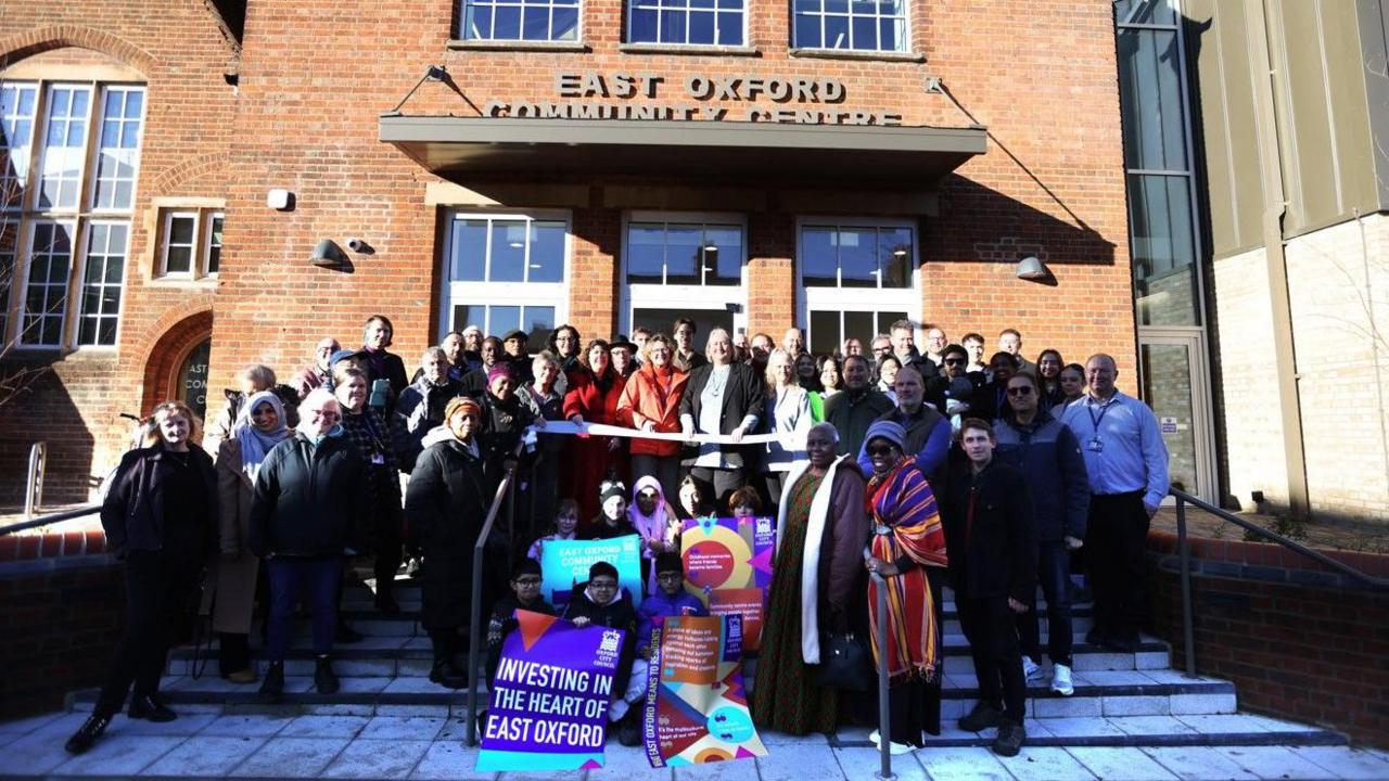 A crowd of smiling people has gathered on the front steps of the East Oxford Community Centre.  A ribbon has been stretched out across the top step and a woman wearing an organge jacket is posing with some scissors.