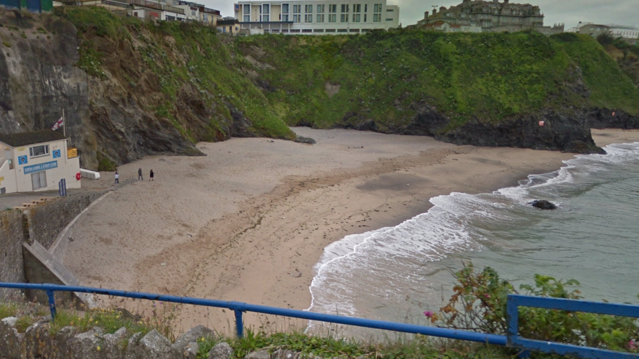 A heightened view looking down on to Great Western beach. It is an overcast day, there are cliffs at the back of the beach which are covered in grass and moss. The tide is halfway out and there are two small figures walking on to the beach. Above the cliffs are a series of buildings. 