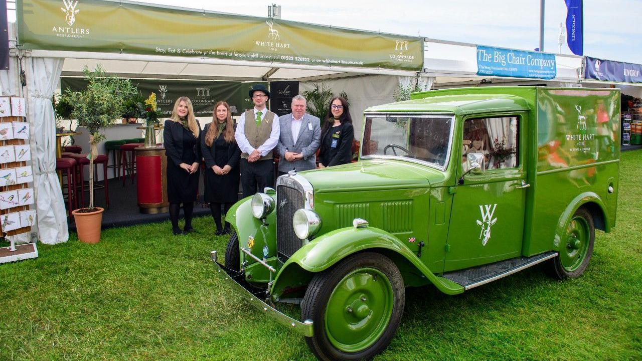 A classic green car with 'White Hart' and a stag logo on the side of it. Behind it a group of people are stood in smart clothes in front of a White Hart trade stand with bar stools dotted around.