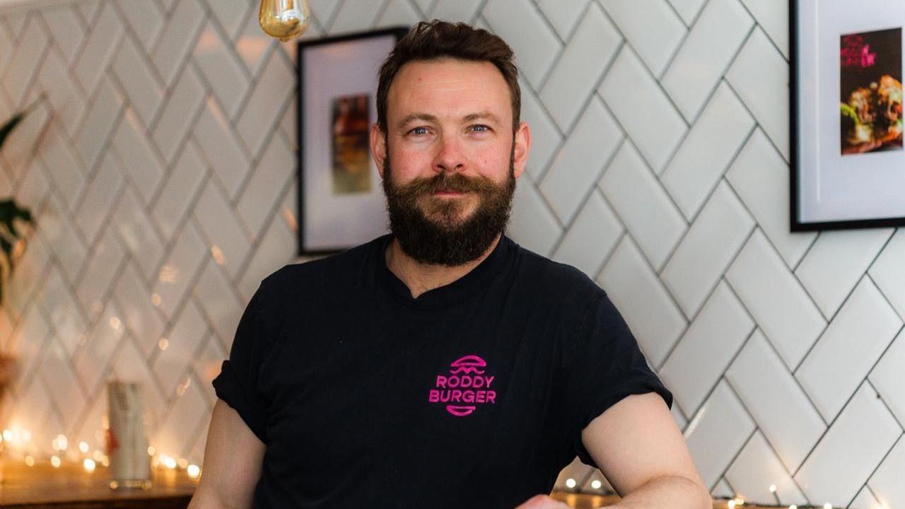 A man with dark hair and a thick beard standing in front of a white tiled wall. He is wearing a black shirt which says "Roddy Burger" on the front in pink text.