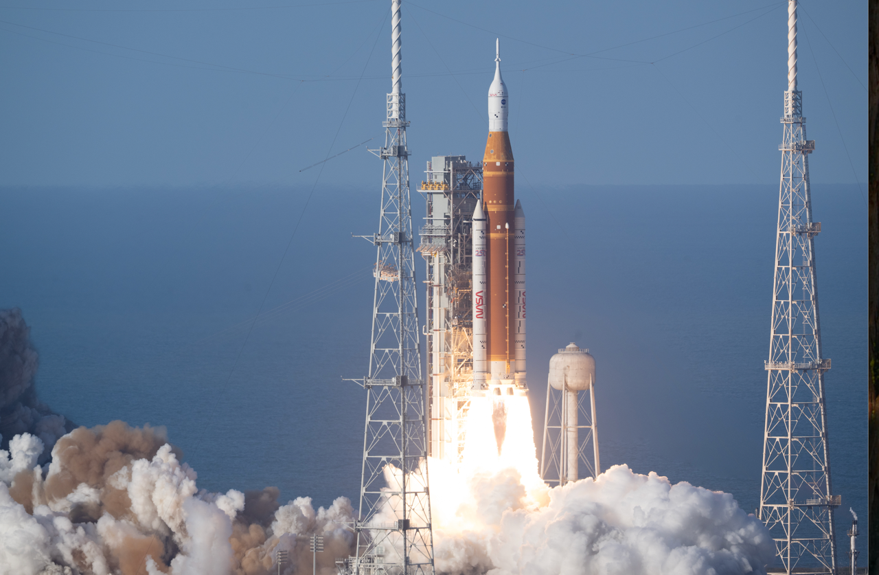 A tall white and orange rocket is blasting off from a coastal launch pad. Flames and bright white exhaust pour from its base, creating a wide, glowing tail that hides the ground in thick clouds of smoke. The rocket is pencil‑thin and points straight up into a clear blue sky, with the sea faintly visible behind it. Slender white booster rockets cling to either side of the central orange core. To the left and right stand two latticework metal towers, like giant scaffolding poles, framing the rocket. A rounded white tank sits nearby on spindly legs, partly lost in the steam. The overall impression is of immense power and light as the vehicle climbs away, leaving a boiling, churning cloud where it stood.