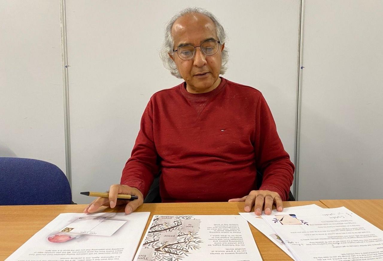 A man wearing a red jumper and glasses is sitting at a wooden table, looking at sheets of paper with writing on them