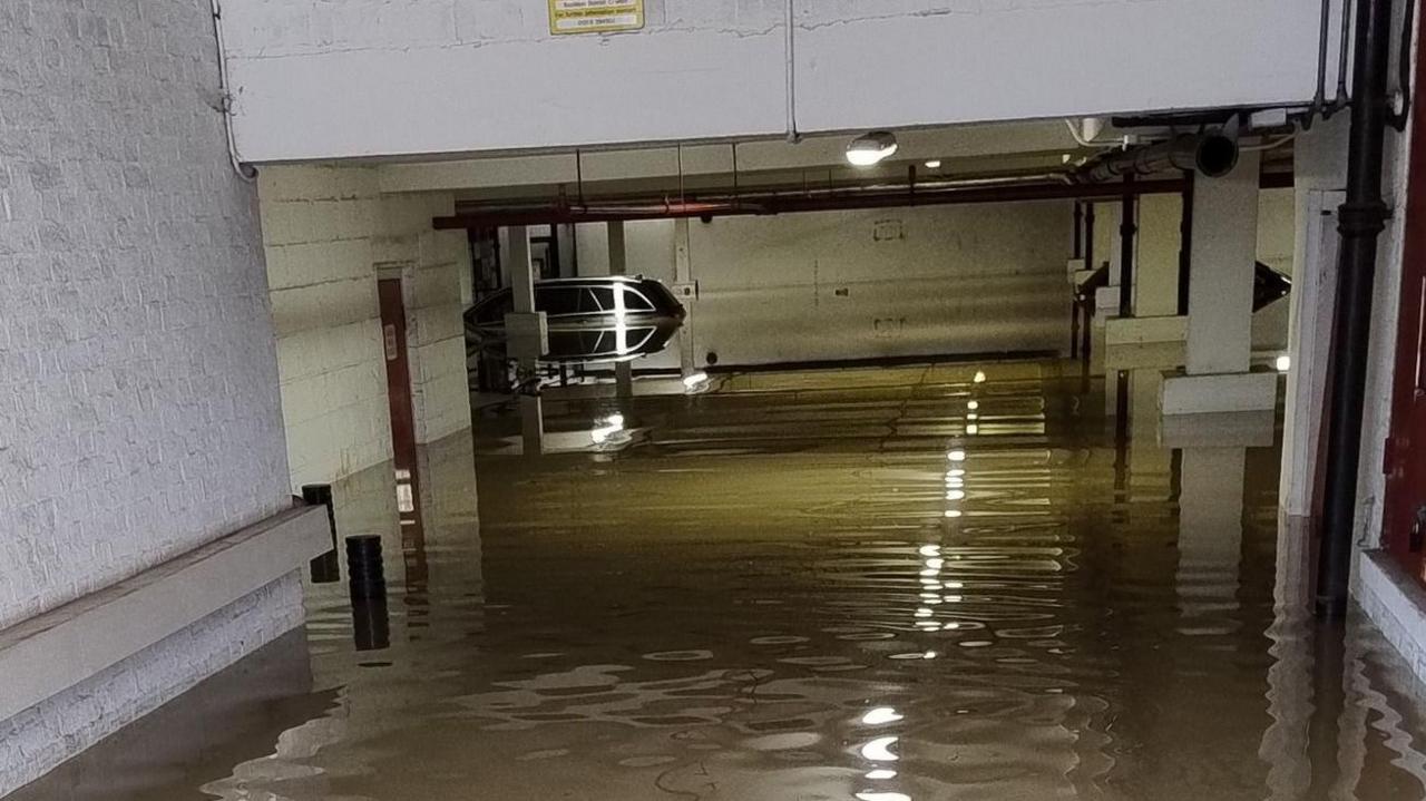 An underground car park is flooded with water. A car is at the back of the image and the water is up to its windows. A light is on in the middle of the ceiling. 