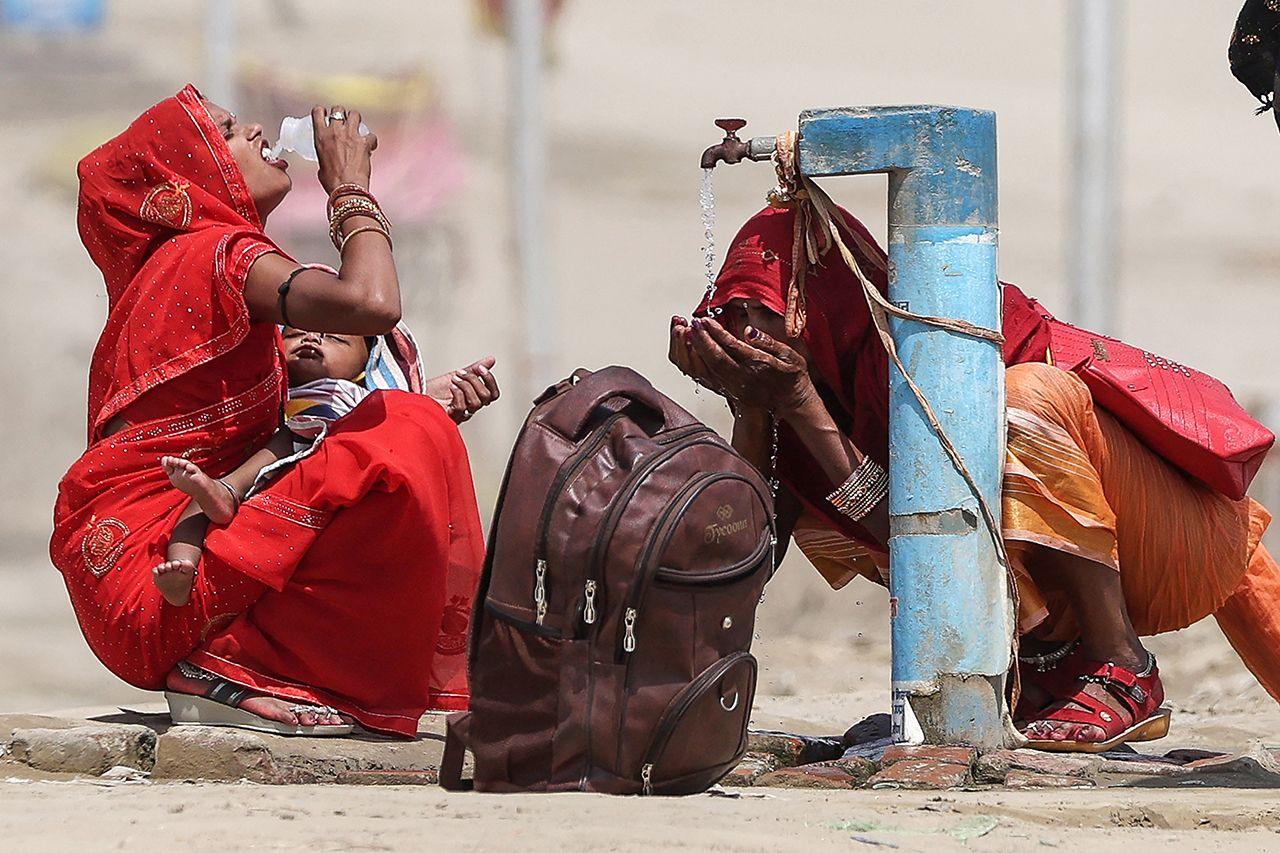 Two women drink water and splash their face from a public tap on a street in Prayagraj, India, on 10 June. They both wear red, with one holding a baby and a backpack on the ground next to them.