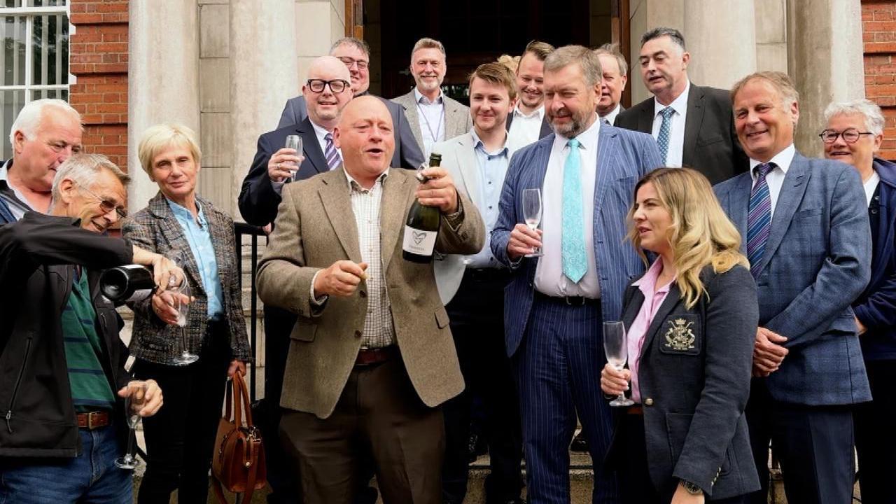A group of men and women, most wearing suits or blazers, hold champagne flutes as a man at the centre uncorks a bottle of champagne. They stand in front of a council office with red-brick walls and a stone doorway.