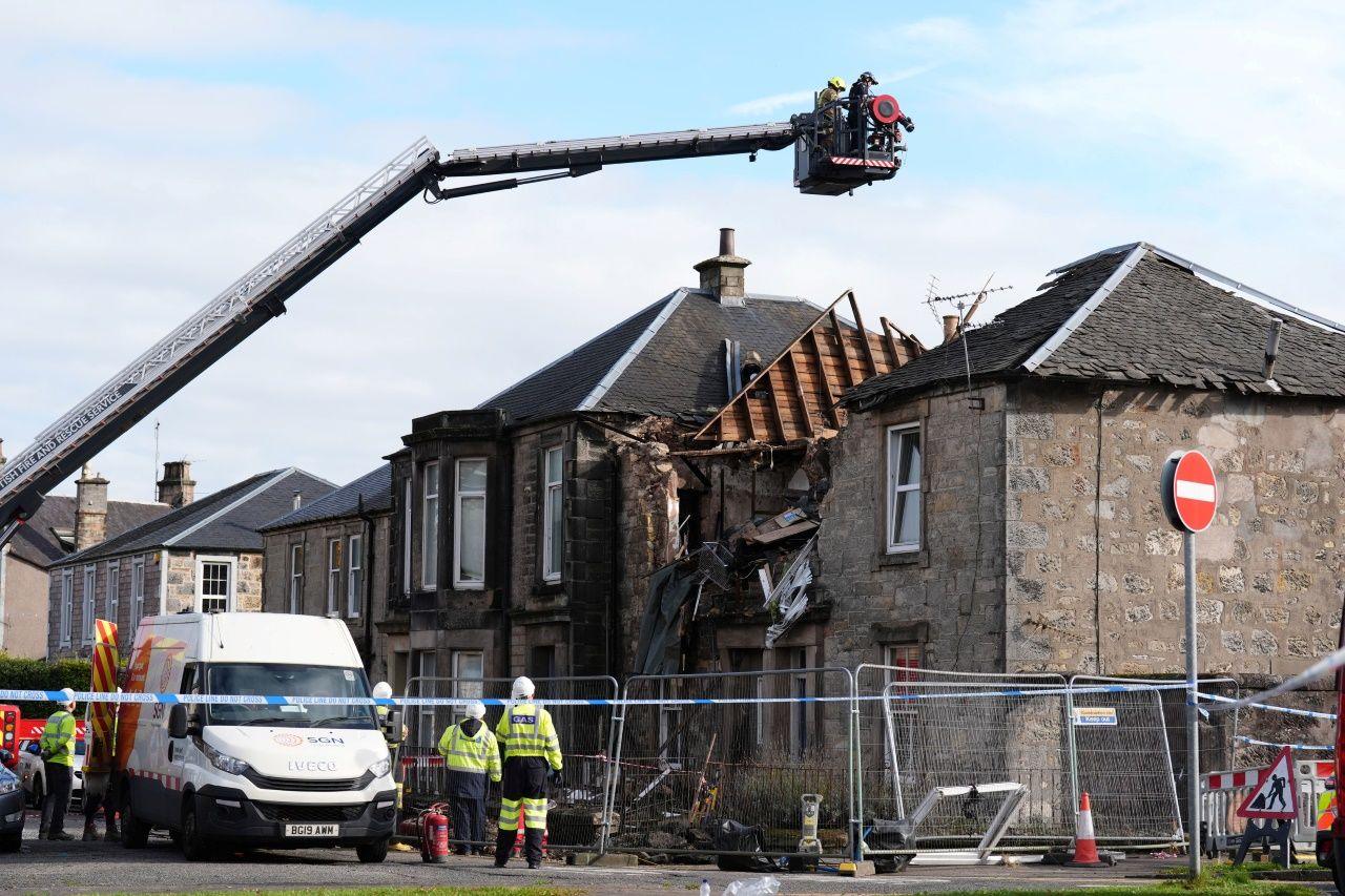 Firefighters on a crane survey the remains of a flat destroyed in an explosion. The foreground is cordoned off and emergency service workers are seen beside the police cordon. 