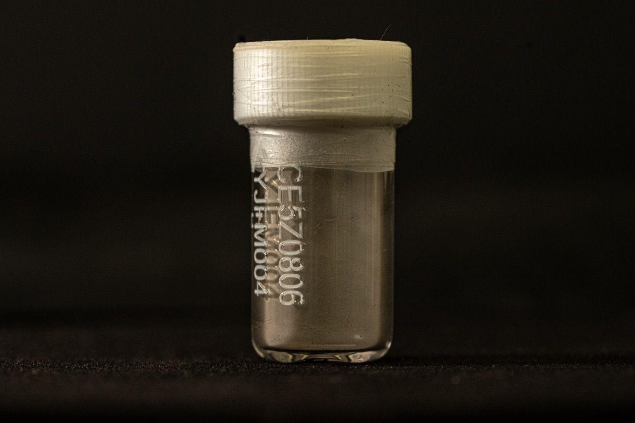 A small clear vial with a white plastic top and tape around the lid. It is lit against a black background. On the front is a code. Inside at the bottom there is a coating of dust.