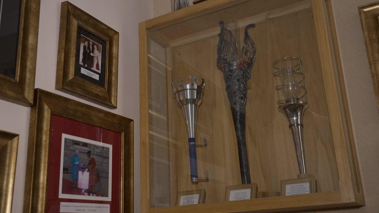 Ceremonial torches in an enclosed glass and wooden-framed case are mounted on a wall. Adjacent are gold-framed photos including one of the late Queen Elizabeth II with a Beefeater in his red and gold outfit.