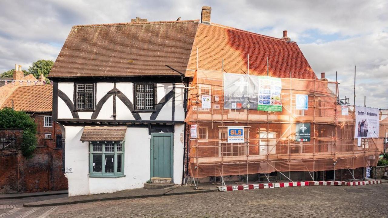 The old property on the left is white with black markings on the first floor and a green window and door on the ground floor. The building on the right has scaffolding and red netting