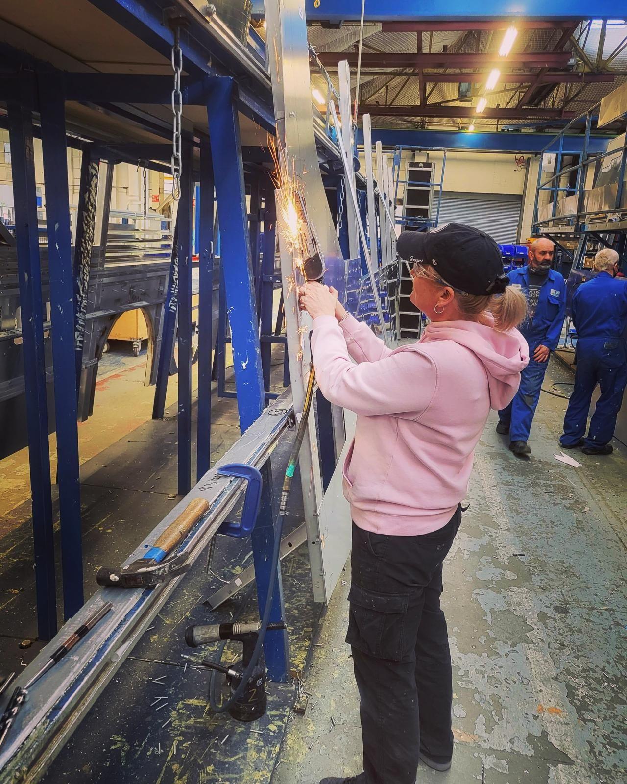 Laura Pollard, in work trousers and a pink hoodie, hat and goggles, cuts a piece of metal on a bus being built in the Dennis factory. Two men in blue overalls are in the background