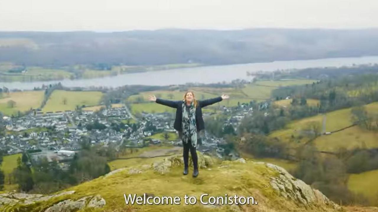 A screenshot of the promotional video featuring a woman standing on the top of a fell with Coniston in the background. The subtitles read "Welcome to Coniston".