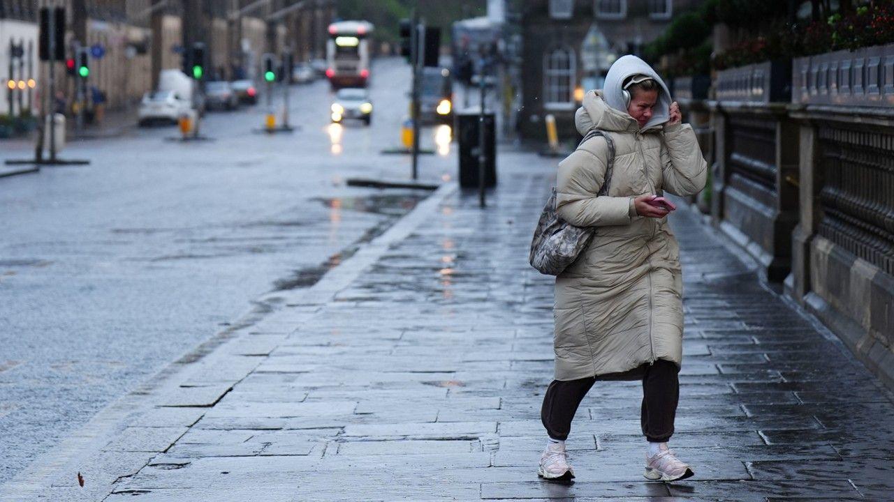 A windswept-looking woman walks down a city street, holding a mobile phone, wearing a long coat and with her hood up. Buses and cars are in the background.