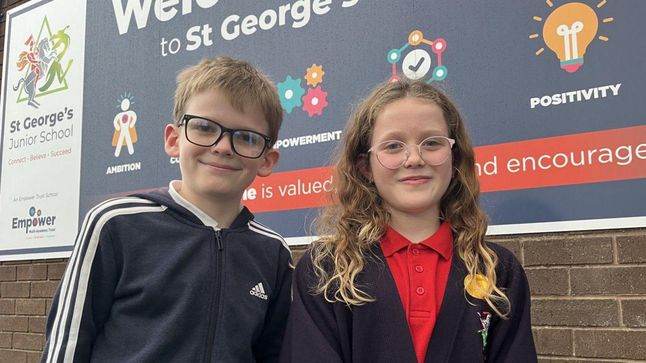 Marcus and Pippa stood side by side, smiling and looking into the camera. Marcus is wearing a navy blue adidas tracksuit top. Pippa is wearing a red polo shirt and a black school cardigan. They're stood in front of a blue sign that reads 'Welcome to St George's' in white writing. 