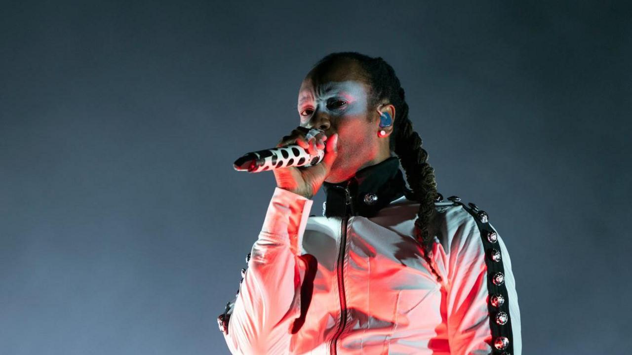 Maxim, with dreadlocks tied in a plait, holds a microphone up to his mouth, wearing a white jacket with a black strip down the arms with bells on.
