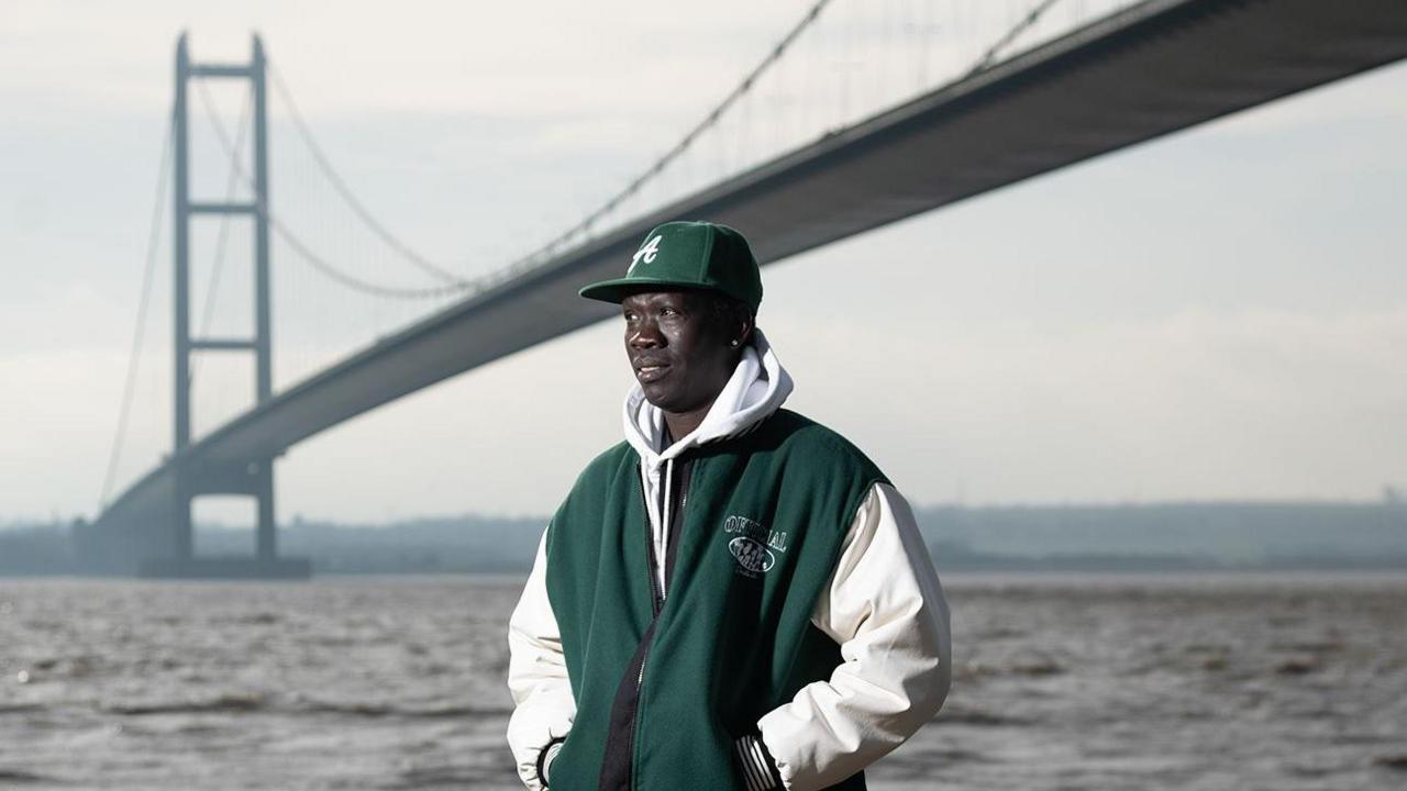 Anei smiles as he looks ahead, with the Humber Bridge in the background. Anei is wearing a bottle green cap and matching hooded jacket, blue jeans and white trainers.