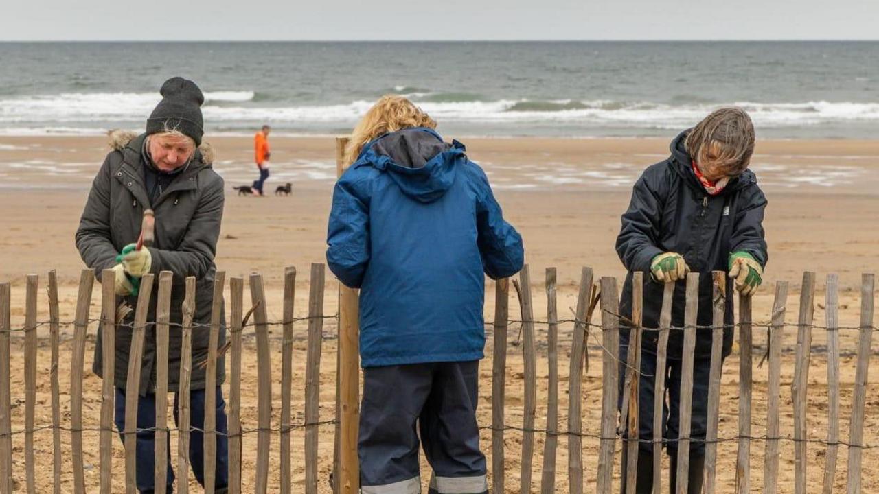 Three volunteers wrapped up with thick coats working on the wooden fence on the beach at Seaton Carew. A man in an orange jacket can be seen walking dogs in the background.
