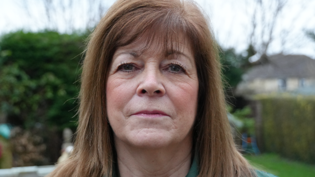 Sue Maggs looking into the camera. She is stood in a garden with bushes and trees visible behind her.