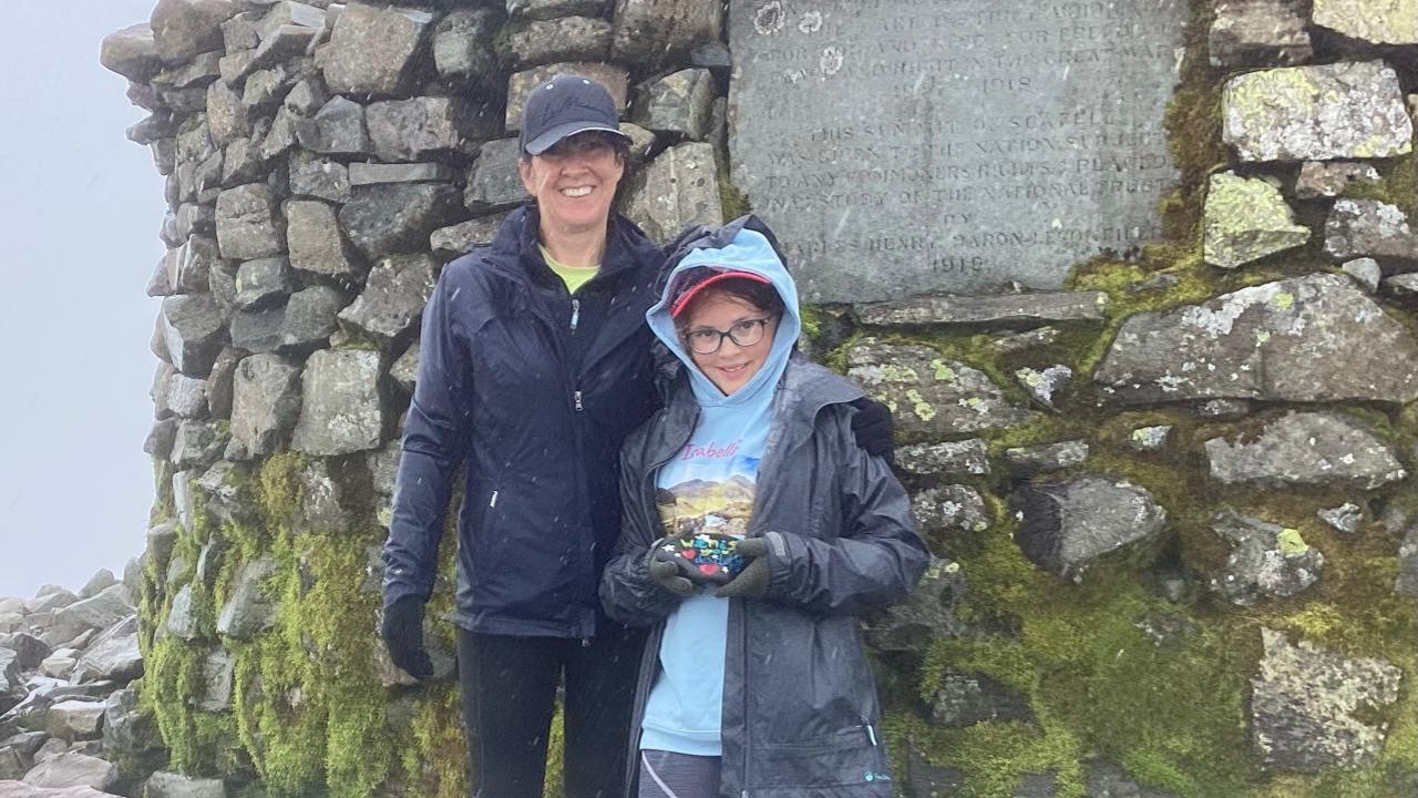 Isabella and her mum standing in front of the trig point on the summit on an inclement day, both wearing rain coats with baseball caps on. 