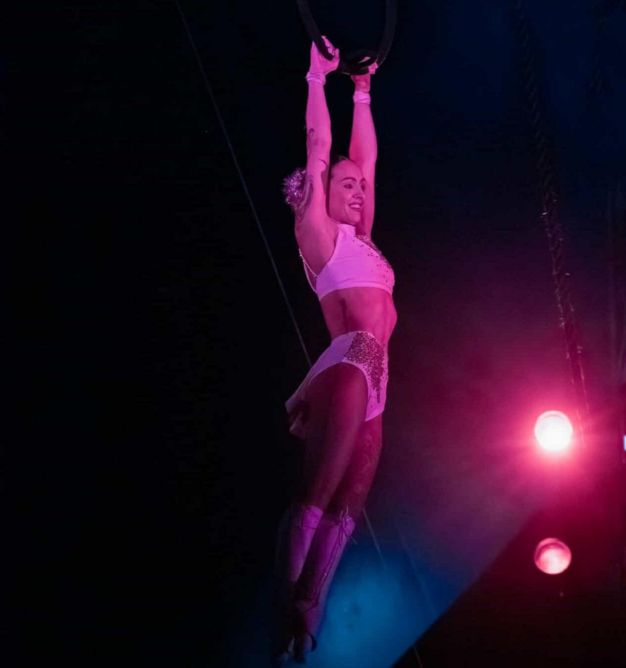 A picture of Kelly performing in the circus. She is wearing a white two piece outfit and is hanging from a hoop which she is gripping with her hands. She is smiling into the crowd.