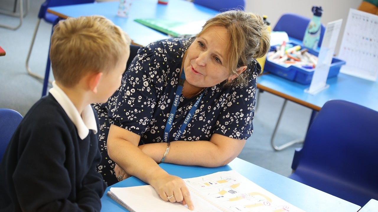 Nicky Coates, who wears a patterned dark dress and a lanyard, sits at a table in a classroom with a pupil. She is pointing at a book while the boy faces her.
