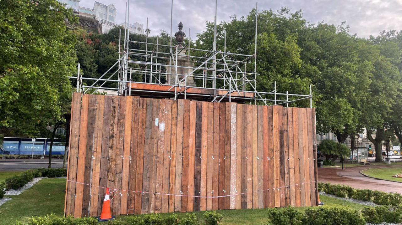 Scaffolding and wooden boards up around Torquay's war memorial. An orange traffic cone is also propped up by the boards and grass and shrubberies surround the memorial.