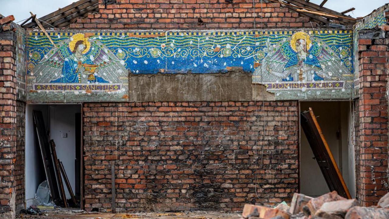 The exposed gable end of one of the convent buildings, which has been partially demolished, and has exposed a mosaic of two angels facing each other from opposite sides of the building.