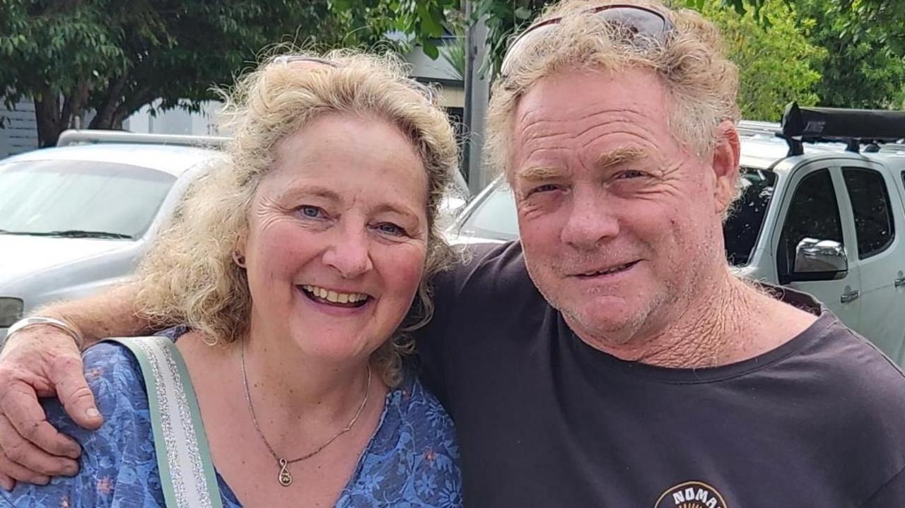 Jess and Alistair, who has his arm around his sister. Jess is wearing a blue top or dress, a necklace and has curly blonde hair. Alistair has a black or navy t-shirt and also has blonde curly hair. They are outside and there are cars parked behind.