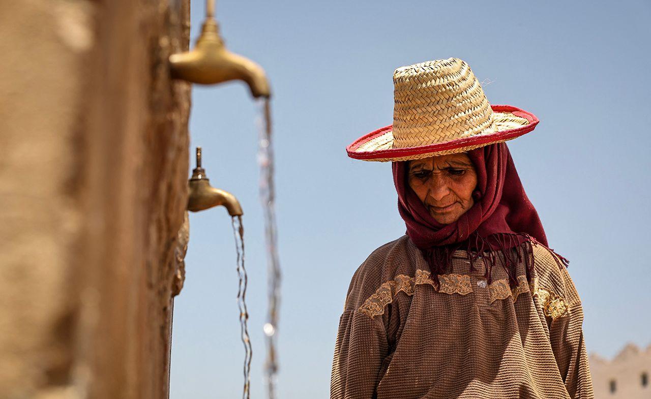 A person looks down while wearing a sunhat with taps pouring water into containers (out of frame) with a blue sky in the background, in Morocco in June 2024