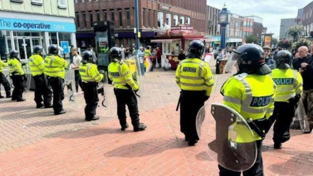 Police officers in a line, wearing riot gear and holding shields, stand near the Blue Clock in Hanley.