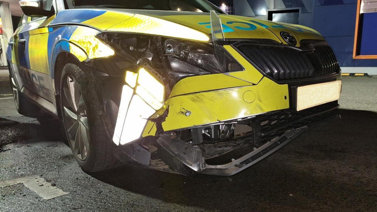 A yellow and blue police car with extensive damage to the front. The headlights are smashed an the bumper is falling off. 