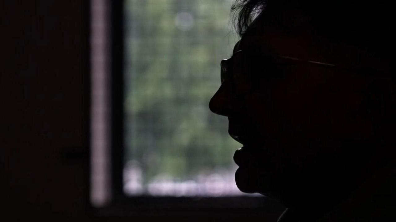 A silhouette of a man's side profile, he has his mouth open as if he is speaking and is wearing glasses. There is a window in the background through which foliage can be seen.