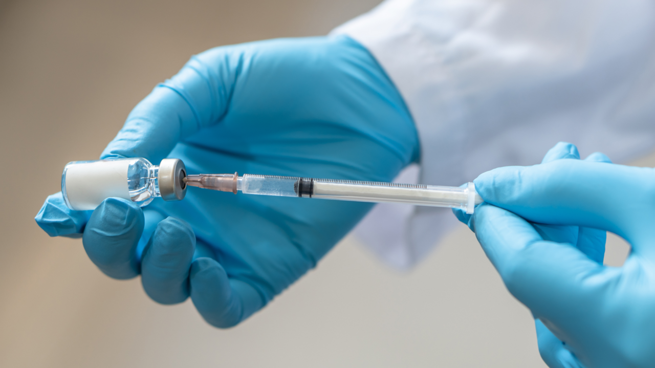 A close up of the hands of a person wearing blue surgical gloves and a white lab coat filling a medical syringe from a bottle.