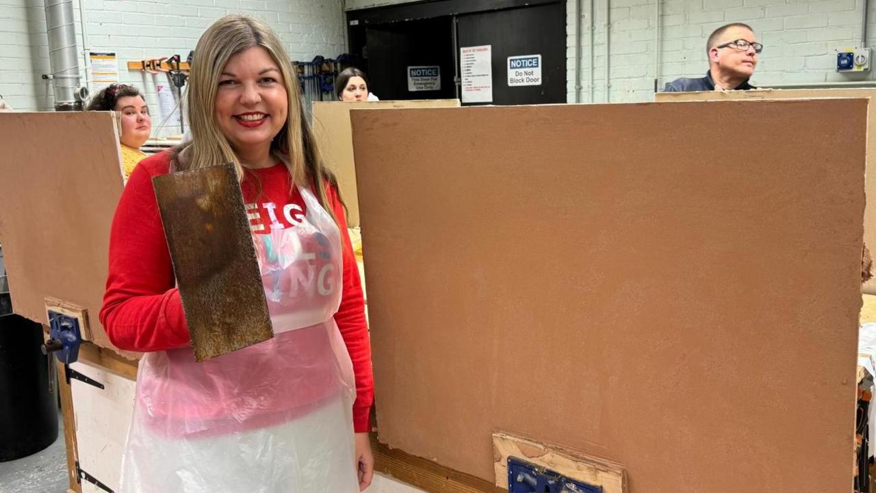 A woman with blonde hair, smiling, wearing a bright red sweater and white plastic apron holds up a plastering trowel next to a freshly plastered board