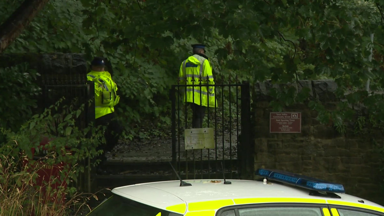 Police officers can be seen inside the gates of Dukinfield Cemetery, with a police car parked on the road near by