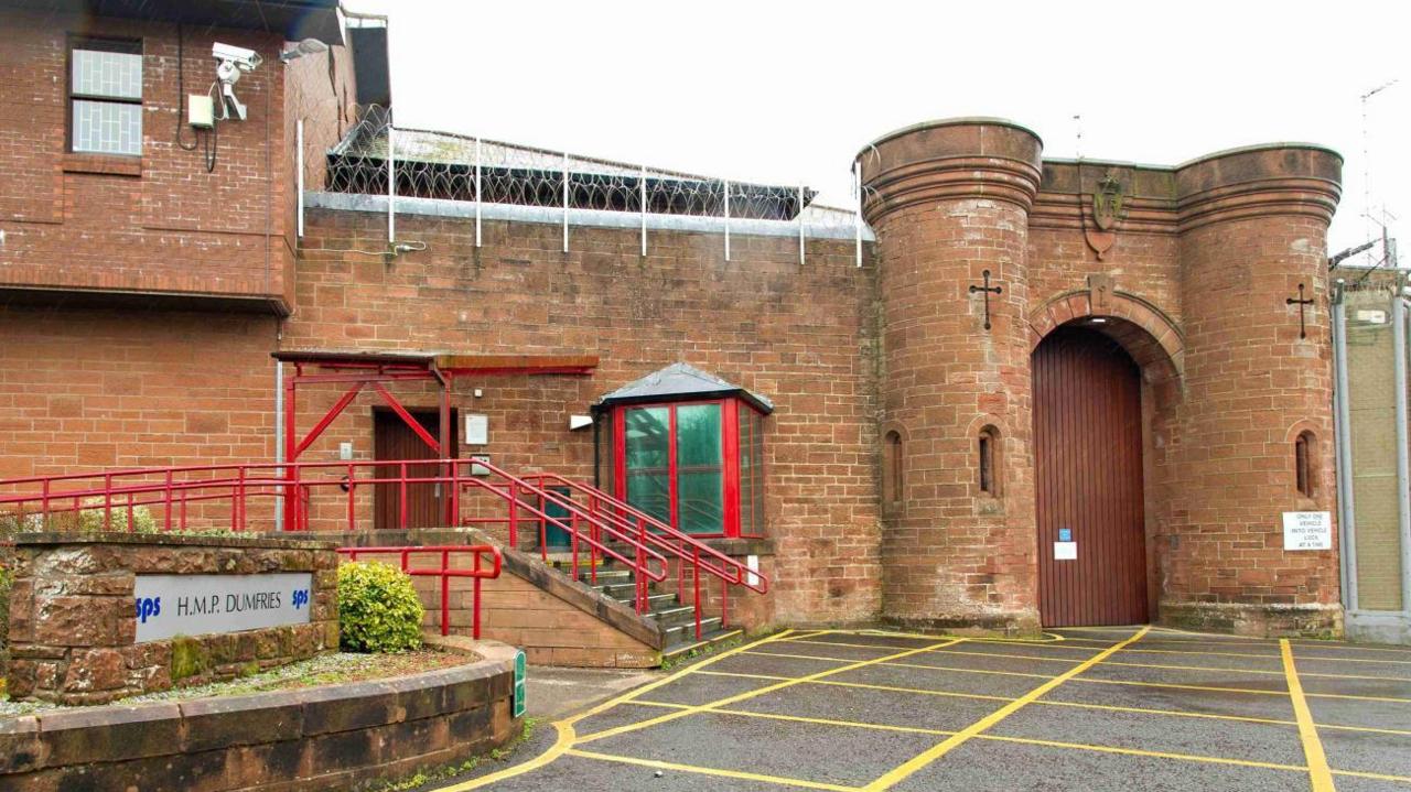 An image showing HMP Dumfries. The building is sandstone, there is a sign to the left stating HMP Dumfries, and red railings on the stairs leading up to a brown door.