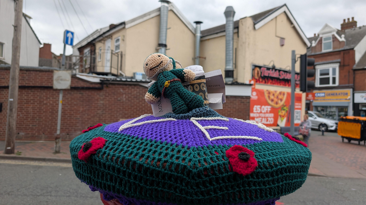 A crocheted postbox topper in green and purple with figure on top, with a high street behind
