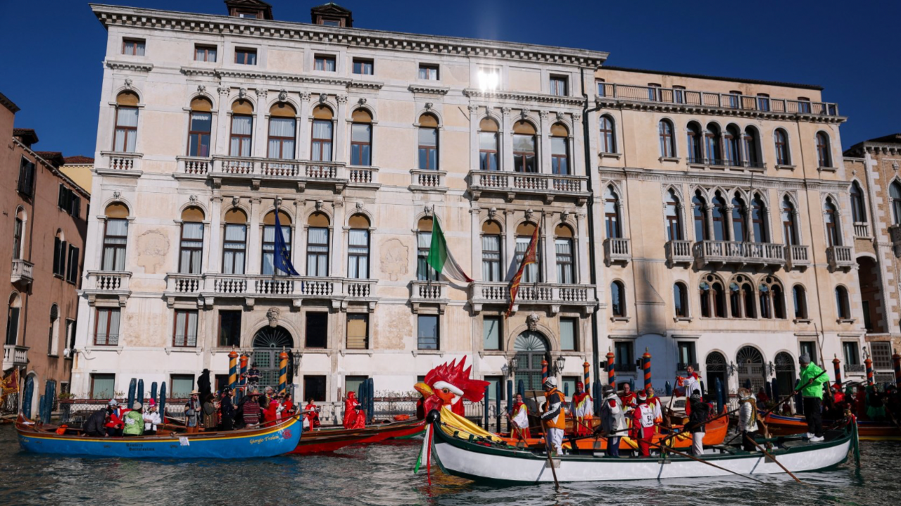 Two boats can be seen on the canal in front of a large building with many windows. One of the boats has the head of a rooster. 