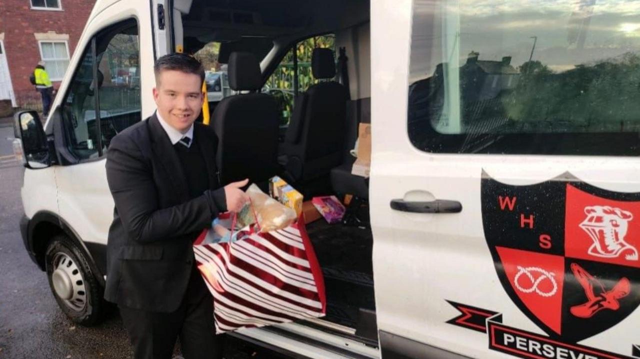 A teenager in a black school uniform holds a red and white striped gift bag with presents in it which he takes out of a white school van with a black and red logo on the door
