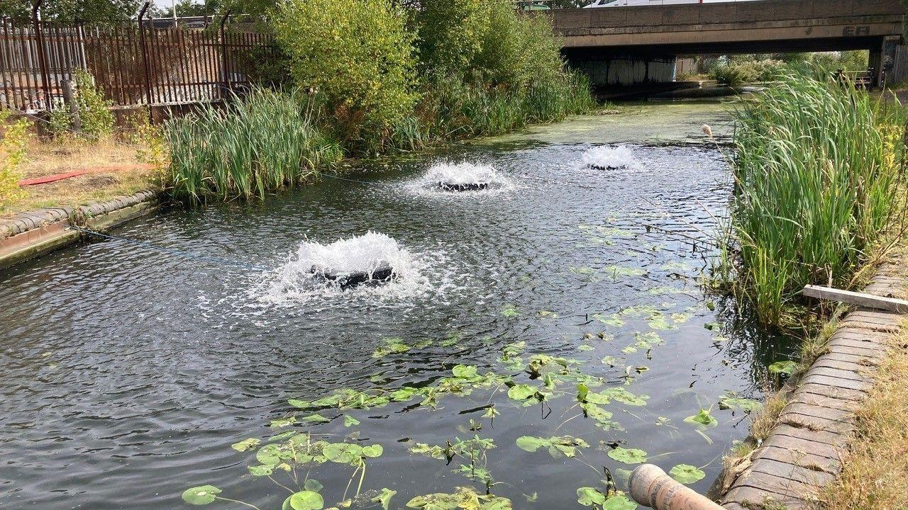 A canal with machines creating bubbles in the surface of the water. there are reeds on either side of the canal, and a bridge in the background